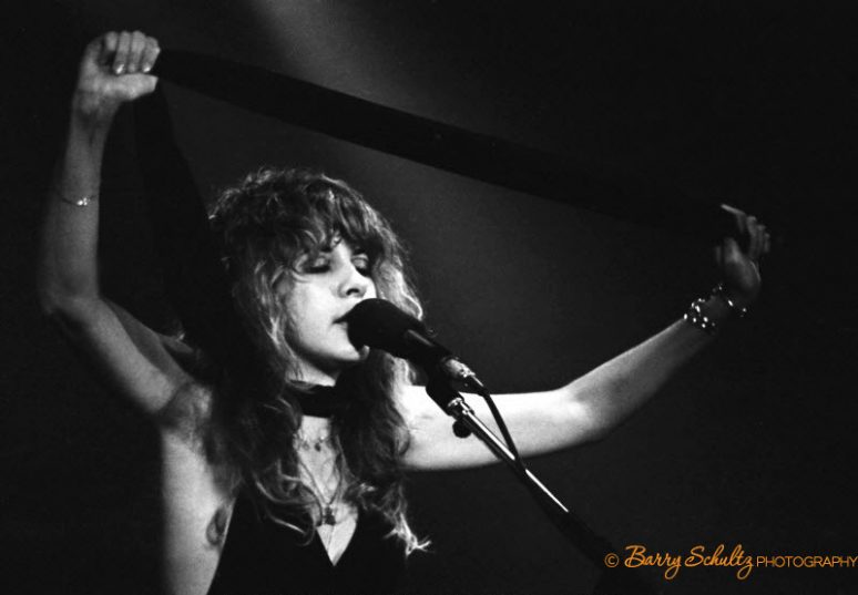 Singer and musician Stevie Nicks of Fleetwood Mac onstage with her arms raised, eyes closed and singing into the microphone.
