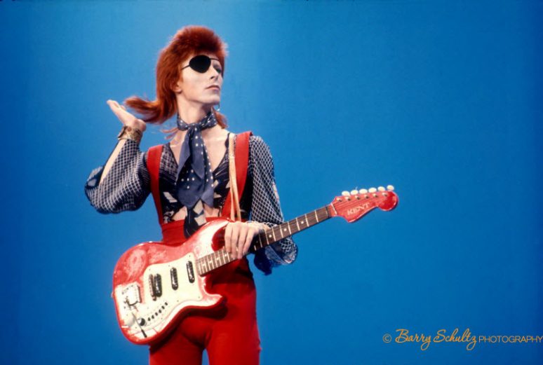David Bowie wears an eyepatch, in red suit with guitar on blue background at TopPop television studios in Hilversum, the Netherlands while doing his hit song Rebel Rebel in 1974.