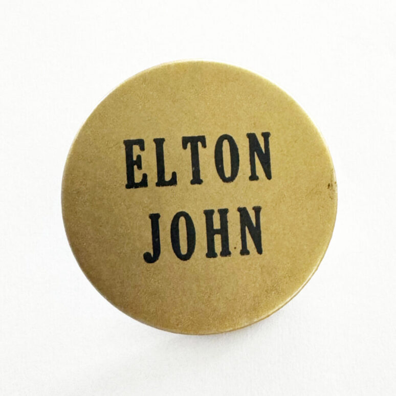 Round gold badge saying 'Elton John' worn when Elton John's Hollywood Star was revealed on the Hollywood Boulevard Walk of Fame in the year 1975.
