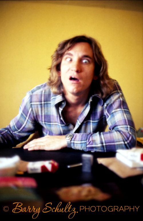 Joes Walsh of the Eagles pulling a face in a blue flannel shirt against a yellow wall.