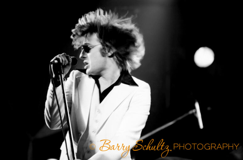 Lead singer of musical group Southside Johnny and the Asbury Jukes, John Lyon, singing live on stage. Wearing a white suit and black shirt with a pointed collar and black sunglasses. He is holding the microphone and stand and flicking his head and hair to the side. Performing in Amsterdam at the venue Paradiso in November of 1977.