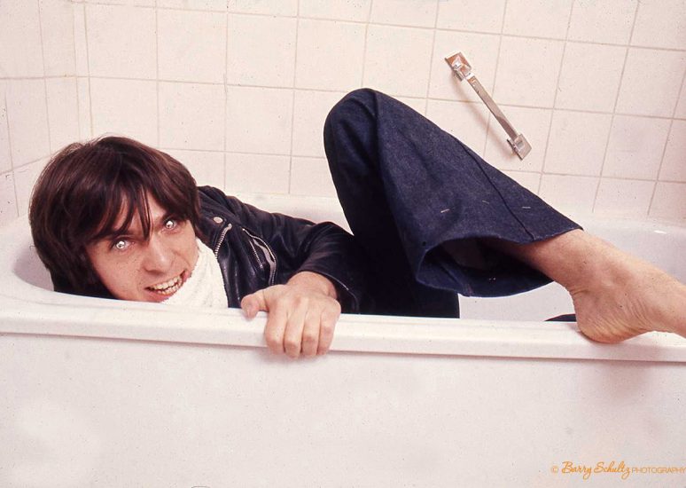 Musician Peter Gabriel posing for a photoshoot with Barry Schultz in his hotel room, lying in his hotel bathtub wearing reflective contact lenses and grinning.