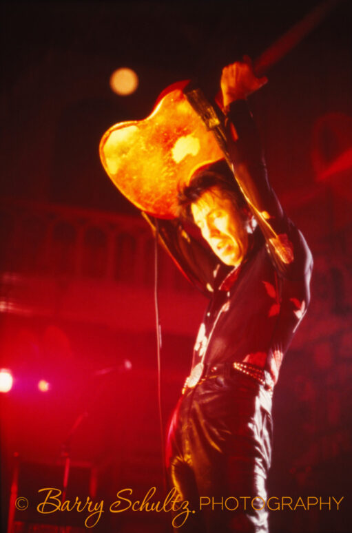 American musician Link Wray lit by a yellow and red light, wearing all black clothing holding his guitar up over his head.