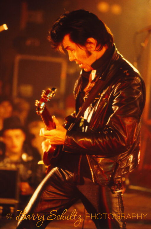 American musician Link Wray lit by a yellow and red light, wearing all black clothing playing his guitar live onstage.