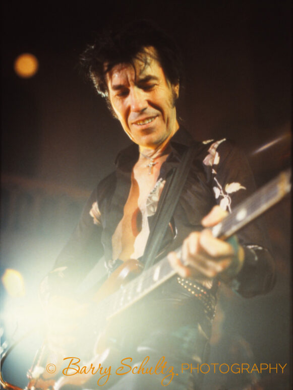 American musician Link Wray wearing all black clothing, smiling and playing his guitar live onstage.
